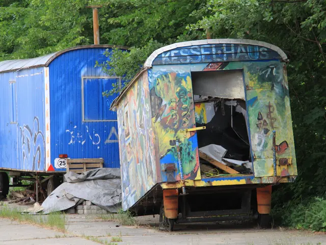 The image shows an old blue trailer with graffiti on it sitting on the side of a road, surrounded...