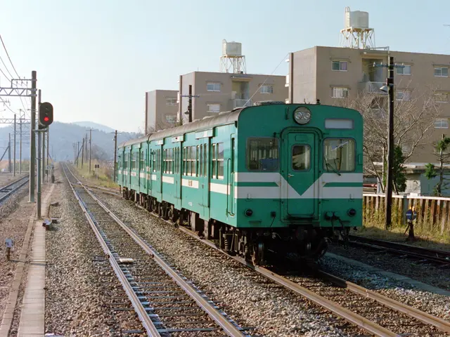 The image shows a green train traveling down train tracks next to a tall building, surrounded by...