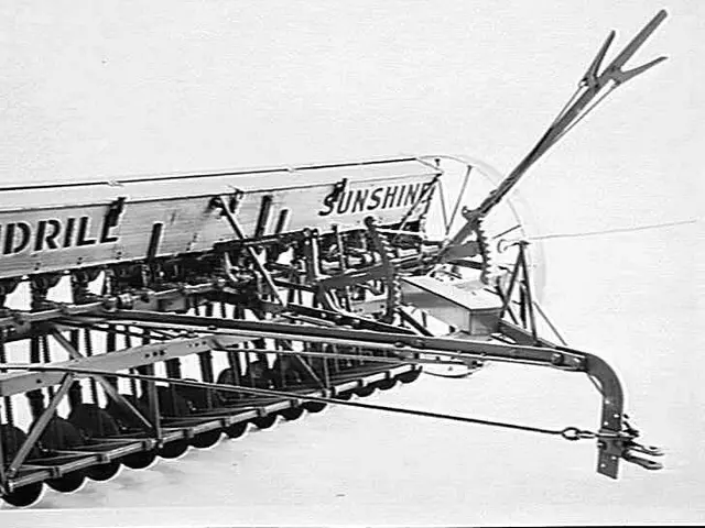 The image shows a black and white photo of a tractor with a seeder attached to it. The seeder is a...