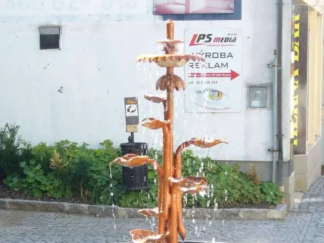 The image shows a fountain in the middle of a cobblestone street, surrounded by lush green plants...