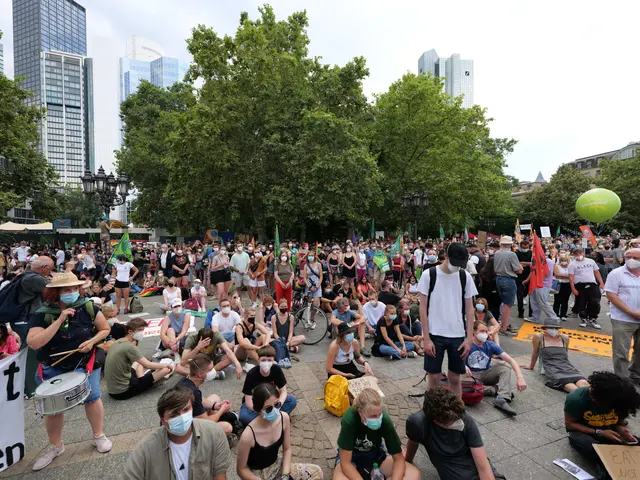 The image shows a large group of people sitting on the ground in front of a crowd of people, some...