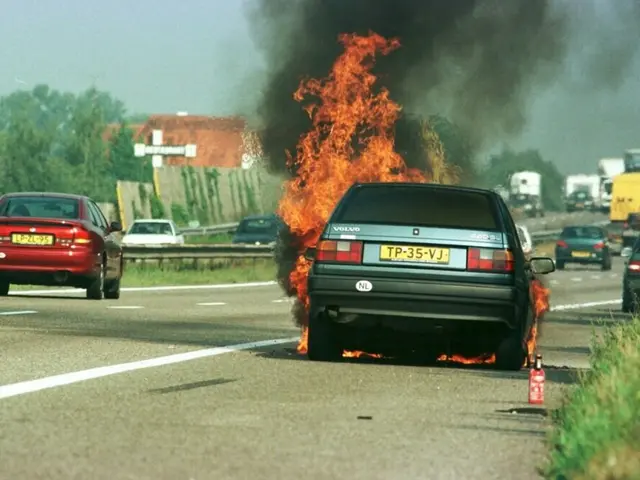 The image shows a car engulfed in flames on the side of the road, surrounded by other vehicles. In...