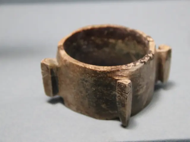 The image shows a close up of a metal ring on display in a museum. The ring is brown in color and...