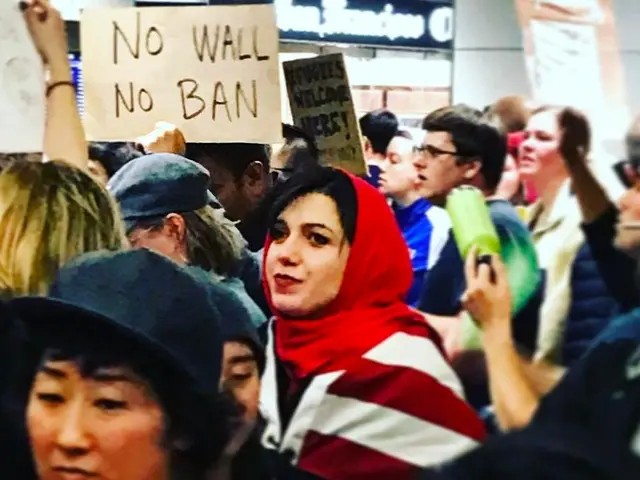 The image shows a group of people standing in front of a building, holding signs that read "No Wall...
