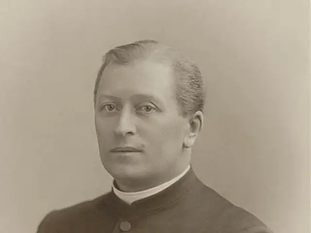 The image shows a black and white photo of a man in a priest's robes, believed to be the first...