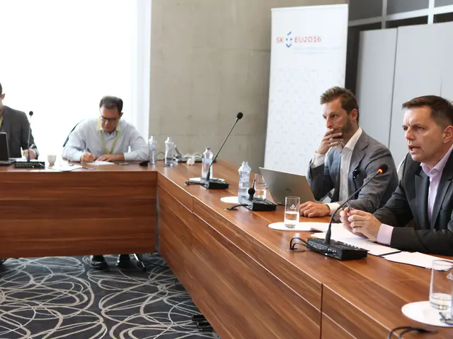 The image shows a group of four men sitting around a long table with laptops, glasses, bottles,...