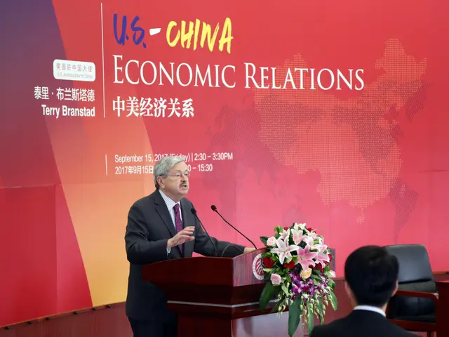 The image shows a man standing at a podium in front of a red wall, wearing a black blazer, white...