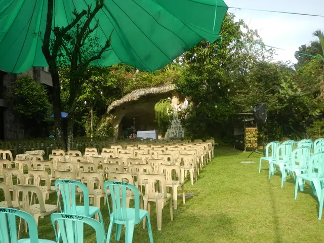 The image shows a wedding ceremony set up in a garden with green chairs and umbrellas, surrounded...