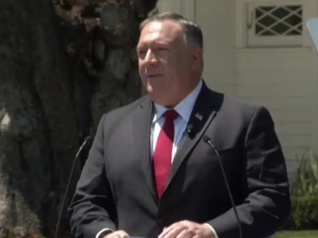 The image shows a man in a suit and tie standing at a podium in front of a tree, surrounded by...