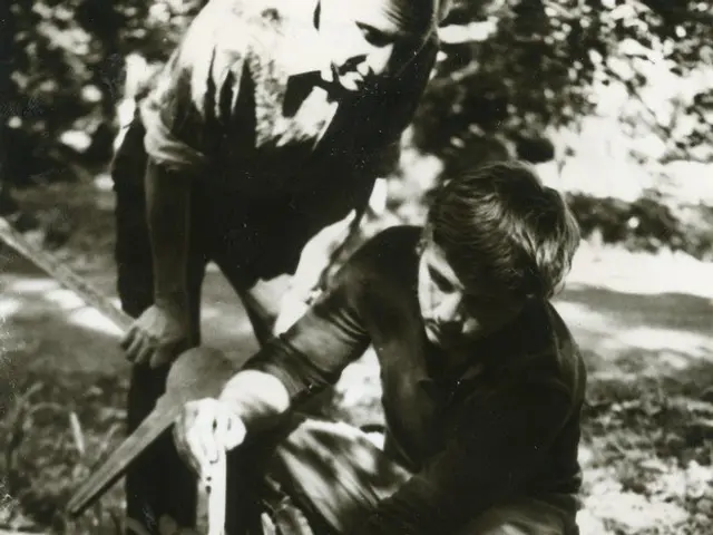The image shows two men working on a log in the woods, with one of them holding a saw in his hand....