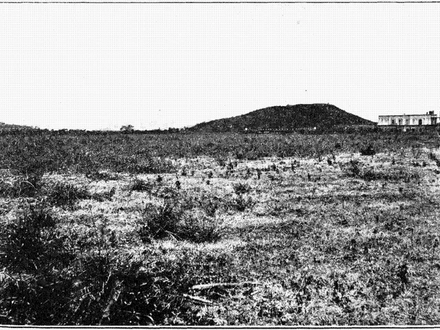 The image shows a black and white photo of a field with a hill in the background, a building on the...