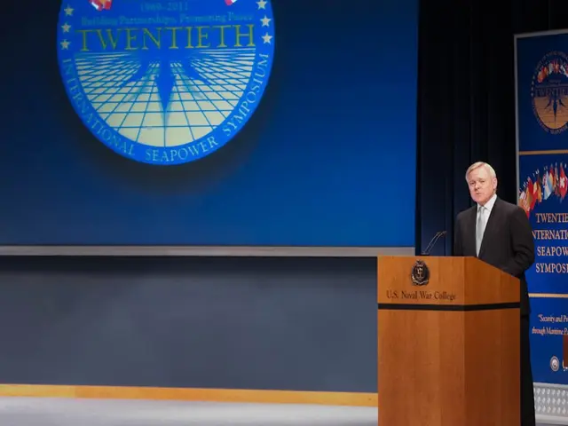 The image shows a man standing at a podium in front of a blue screen, wearing a black blazer, white...
