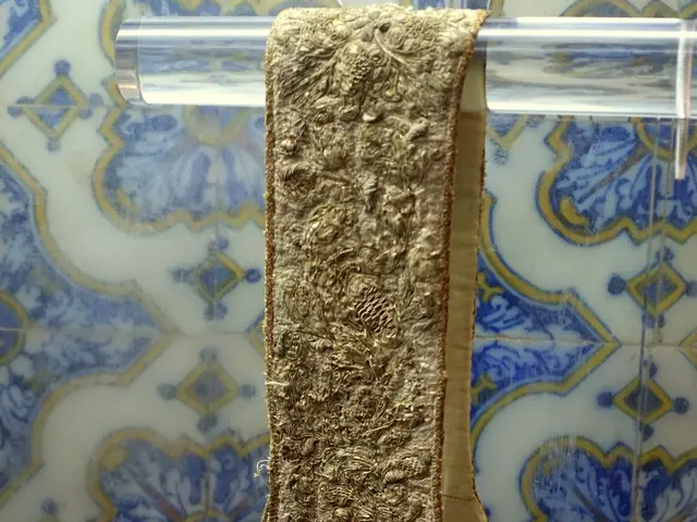 The image shows a gold stole hanging on a wall in a glass case. The stole is intricately designed...