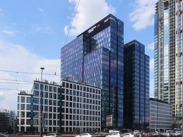 The image shows a large building with many windows in the middle of a city, surrounded by street...
