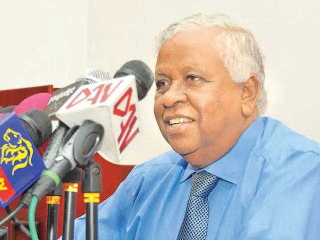 The image shows a man in a blue shirt and tie sitting at a table with microphones in front of him,...