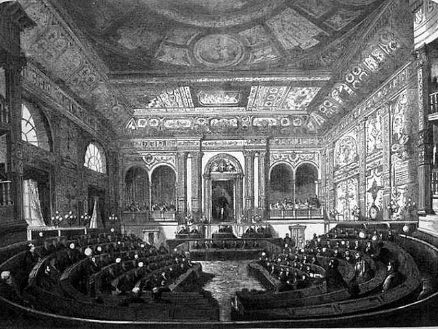 The image shows a black and white drawing of a large auditorium filled with people sitting in...