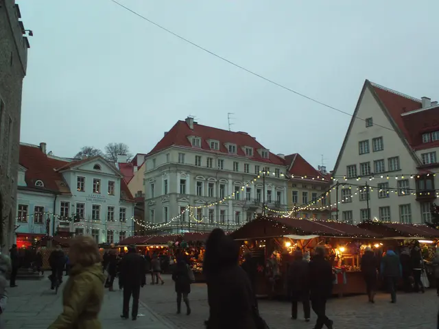 The image shows a bustling Christmas market in Tallinn, Estonia. There are many people walking...