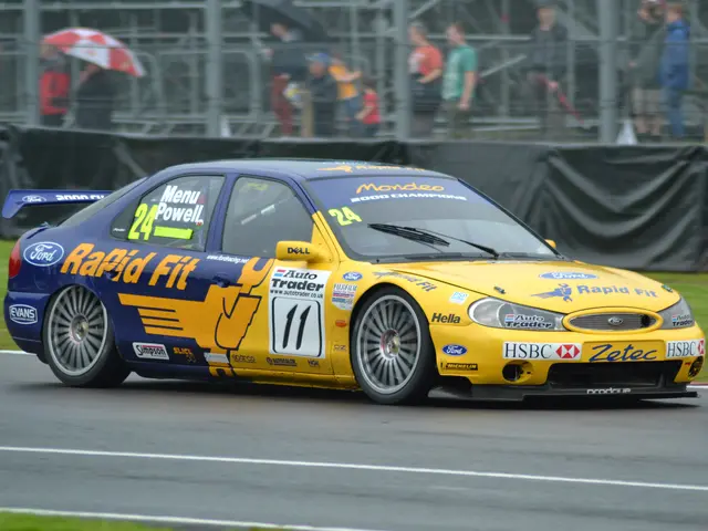 The image shows a yellow car driving down a race track with text and numbers on it, surrounded by...