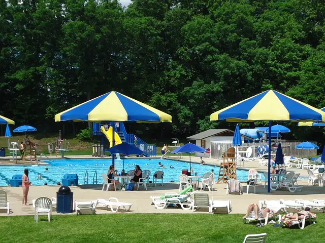 The image shows a large swimming pool surrounded by lounge chairs and umbrellas, with people...