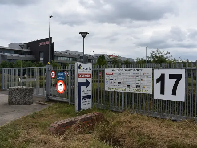 The image shows a large building with a sign in front of it, surrounded by a metal fence, grass, a...