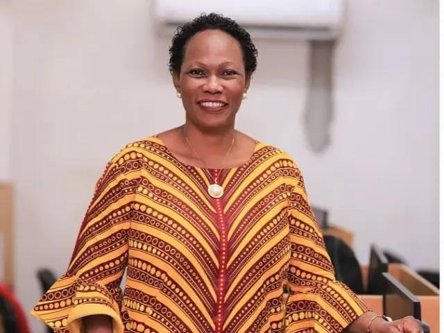 The image shows a woman standing at a podium in front of a microphone, wearing a yellow and red...