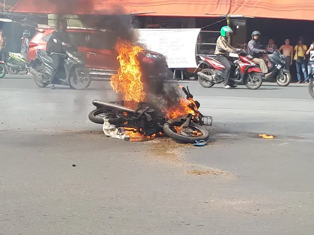 The image shows a motorcycle engulfed in flames on the side of the road, with a car and a van in...