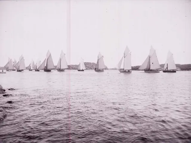 The image shows a black and white photo of a group of sailboats sailing on the water, with hills in...