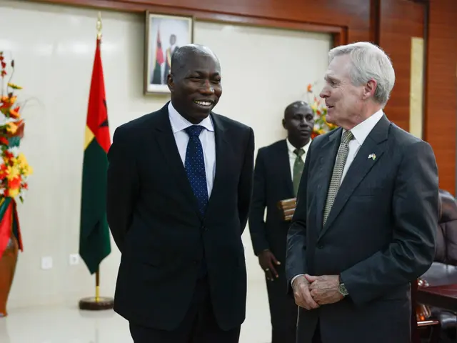 The image shows two men in suits and ties standing next to each other in a room with chairs, a...