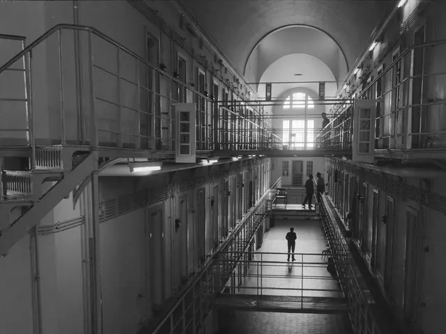 The image shows a black and white photo of a man walking down a long hallway in a prison cellblock....