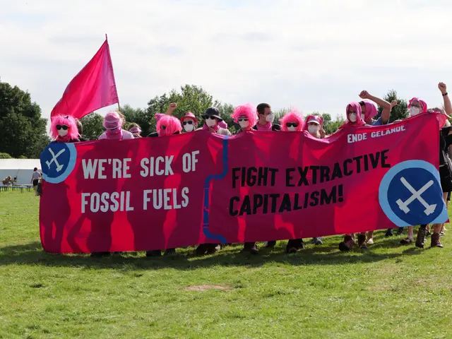 The image shows a group of people standing on a grassy field, holding a pink banner that reads...