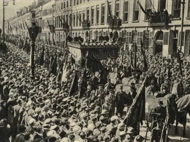 The image shows a large crowd of people walking down a street, with some of them holding flags. In...