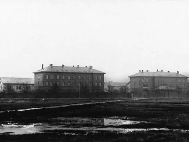The image shows a black and white photo of a large building in the middle of a field, surrounded by...