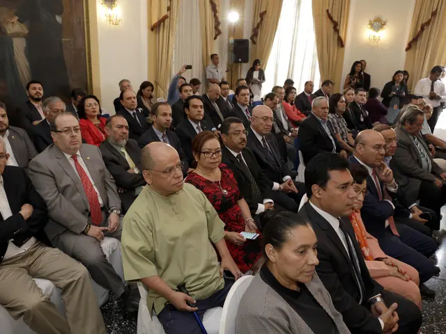 The image shows a large group of people sitting in chairs in a room with a wall adorned with a...