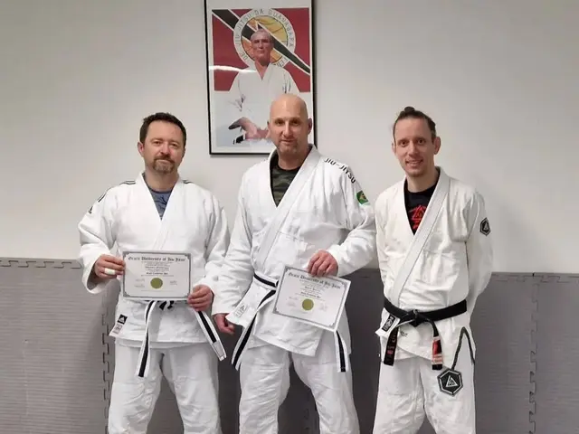 The image shows three men standing next to each other in a gym, all wearing white judo uniforms and...