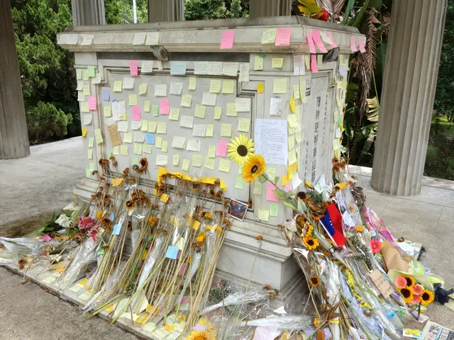 The image shows a memorial with a pillar in the center, adorned with post-it notes and flowers....