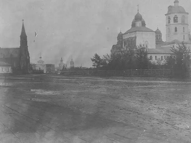 The image shows a black and white photo of a city street with buildings, trees, and a fence in the...