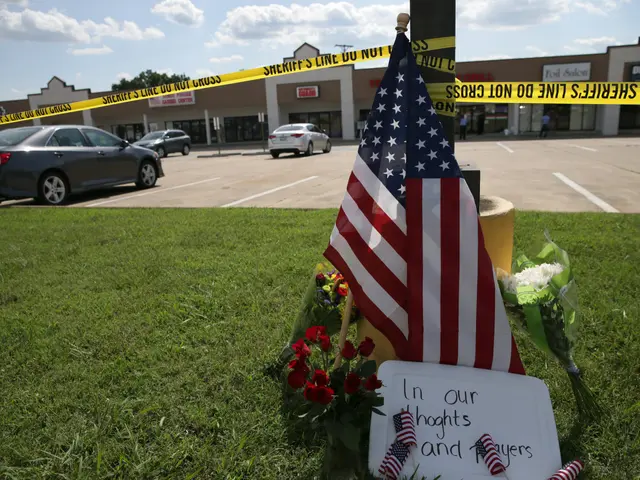 The image shows a memorial in front of a shopping center with an American flag and flowers placed...
