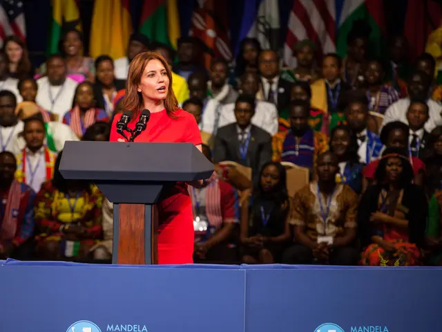 The image shows a woman standing at a podium in front of a crowd of people, with microphones on the...