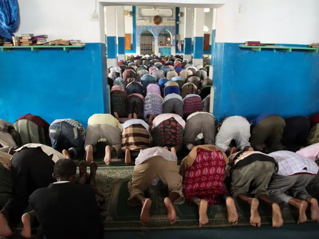 The image shows a group of people praying in a mosque, with books and other objects on the tables...