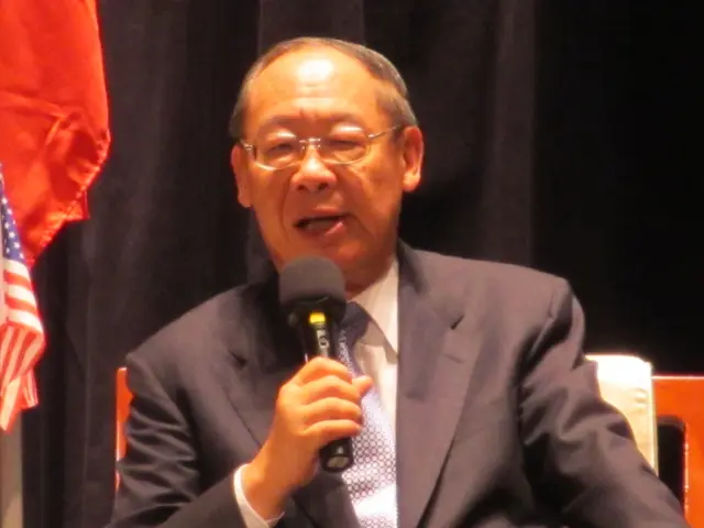 The image shows a man in a suit and tie sitting on a chair, holding a microphone in his hand and...