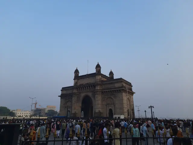 The image shows a crowd of people standing in front of the Gateway of India in Mumbai, India. The...