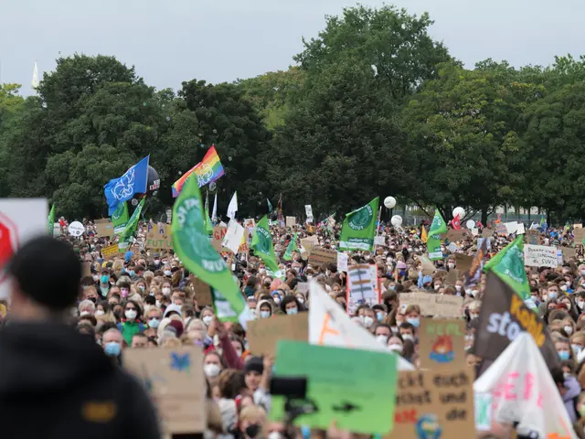 The image shows a large crowd of people holding signs and flags in front of a building, with trees...
