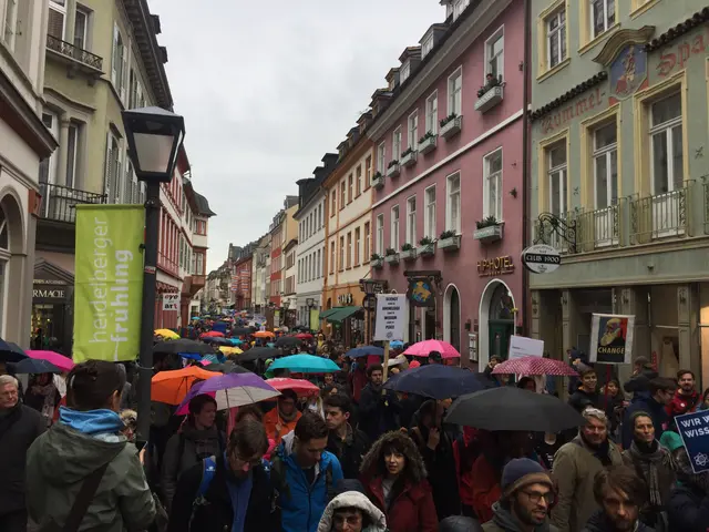 The image shows a large group of people walking down a street with umbrellas, some of them carrying...