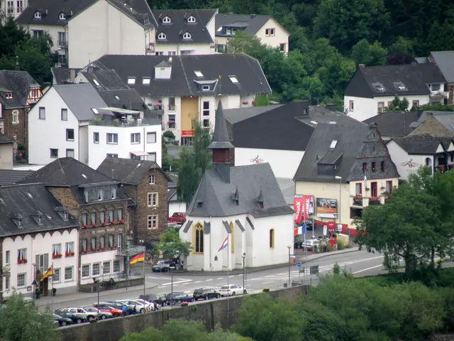 The image shows a small town with many buildings, windows, trees, vehicles on the road, poles,...