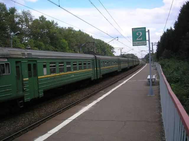 The image shows a green passenger train traveling down train tracks next to a platform, with a...