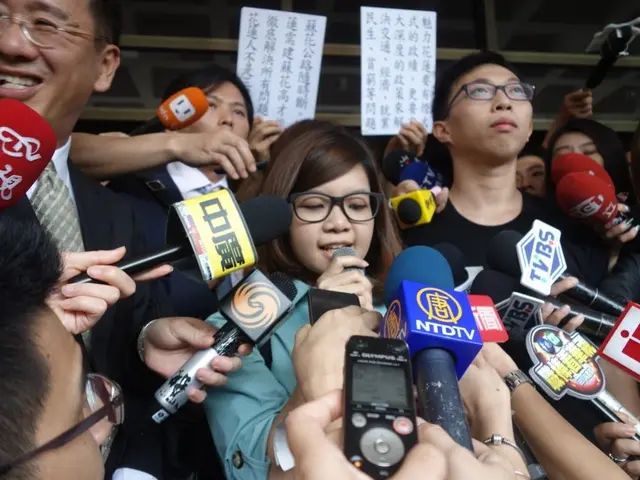 The image shows a group of people standing in front of a crowd of reporters, some of whom are...