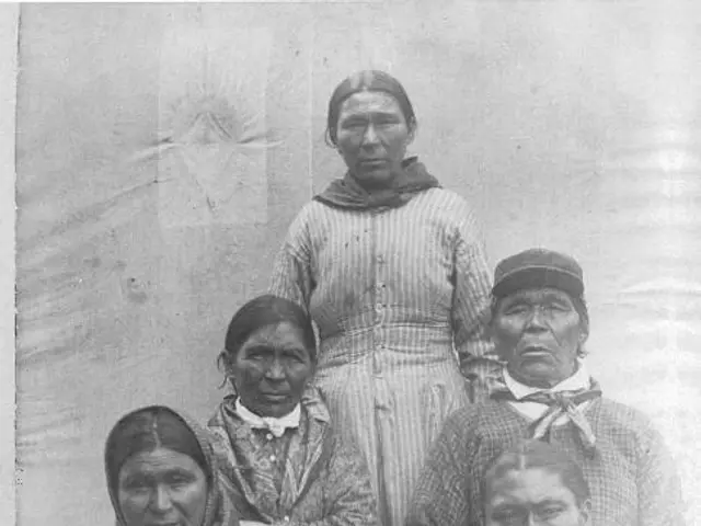 The image shows an old black and white photo of a group of Native American women standing next to...