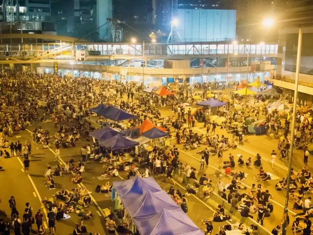 The image shows a large crowd of people standing in the middle of a street at night, illuminated by...