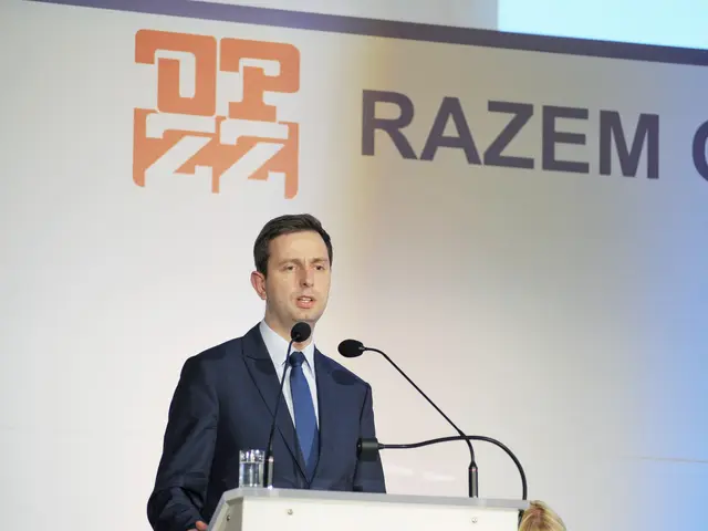 The image shows a man in a suit and tie standing at a podium with a glass of water and two...