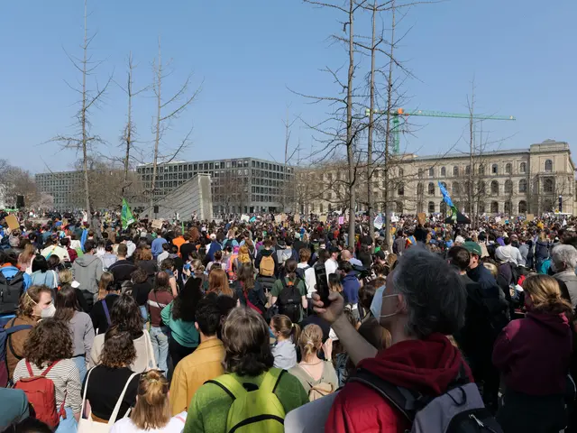 The image shows a large crowd of people standing in front of a building with windows, trees, and a...